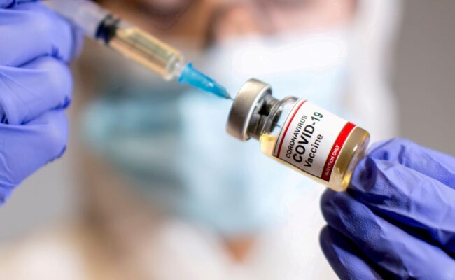 A woman holds a medical syringe and a small bottle labelled “Coronavirus COVID-19 Vaccine